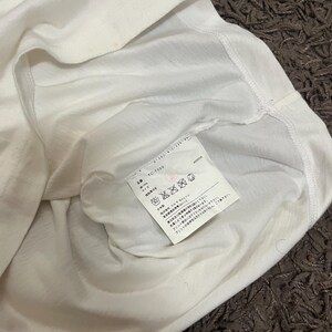 May include: A white t-shirt with a tag attached. The tag has Japanese text and symbols. The shirt is folded, showing the inside of the garment. The fabric appears to be soft and lightweight.