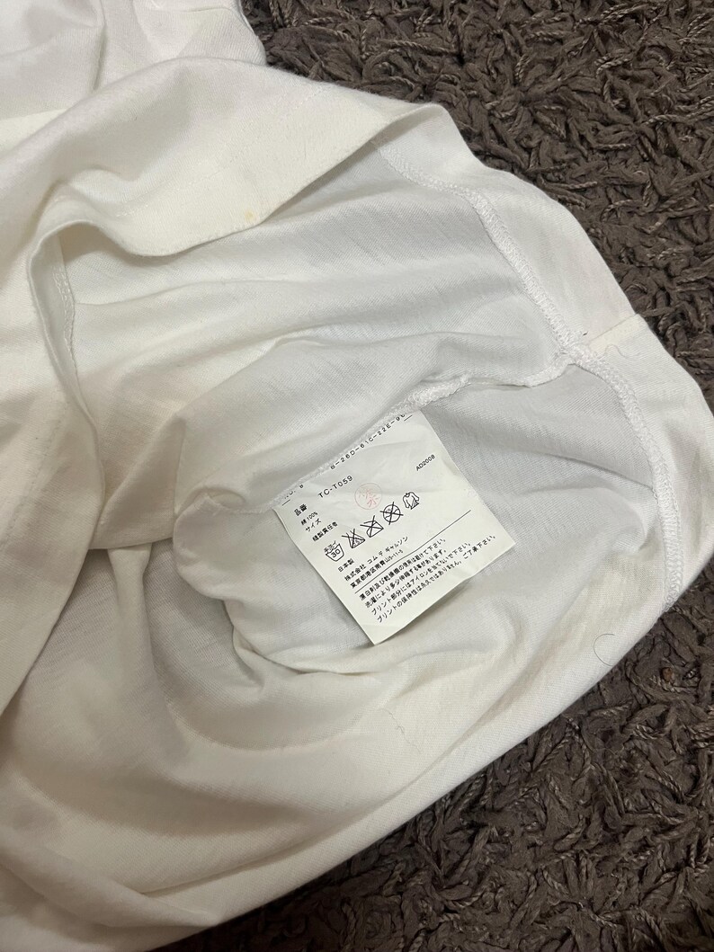 May include: A white t-shirt with a tag attached. The tag has Japanese text and symbols. The shirt is folded, showing the inside of the garment. The fabric appears to be soft and lightweight.
