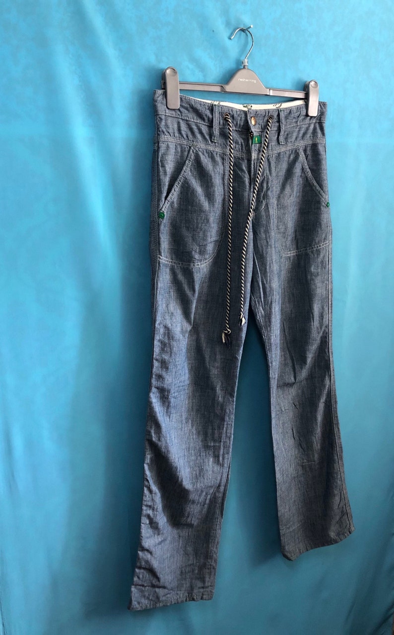 May include: A pair of blue denim trousers with a drawstring waist and two front pockets. The trousers have a subtle striped pattern and are hanging on a black hanger.