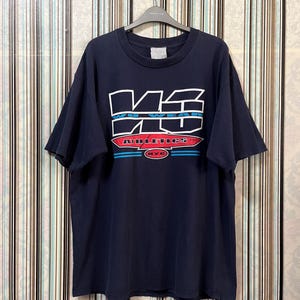 Puede incluir: Camiseta azul marino con un diseño gráfico blanco y azul. El gráfico incluye el texto "WEAR ATHLETICS NYC" en una fuente estilizada. La camiseta está colgada en una percha negra sobre un fondo a rayas.