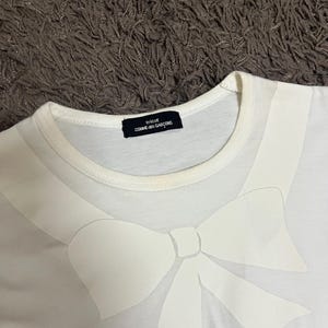 May include: White t-shirt featuring a large, stylized white bow design across the chest. The shirt has a crew neck and a small black label that reads "tricot COMME des GAR&Ccedil;ONS". The fabric appears to be a soft, lightweight material.