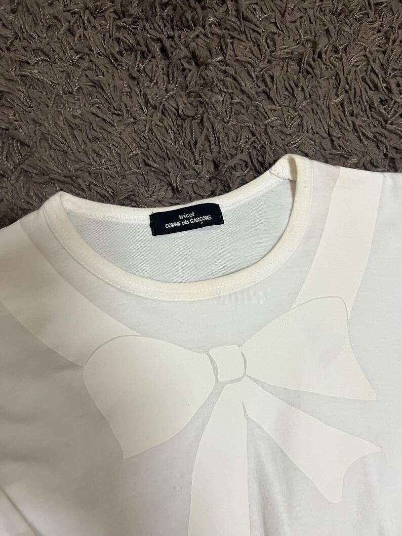 May include: White t-shirt featuring a large, stylized white bow design across the chest. The shirt has a crew neck and a small black label that reads "tricot COMME des GAR&Ccedil;ONS". The fabric appears to be a soft, lightweight material.