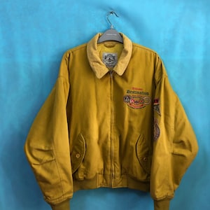 Puede incluir: Una chaqueta bomber de color amarillo mostaza con cuello de pana y cierre de cremallera. La chaqueta tiene un gran gráfico en la parte delantera que dice "Destino desconocido" y presenta una variedad de parches.
