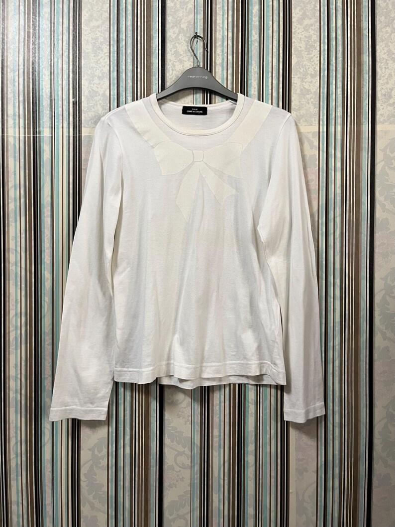 May include: White long-sleeve shirt with a large bow detail on the chest. The shirt has a crew neck and is hanging on a black hanger. The shirt is made of a soft, lightweight fabric.