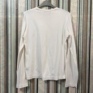 May include: A white long-sleeve shirt hanging on a black hanger. The shirt has a crew neck and is made of a soft, lightweight fabric. The sleeves are long and end at the wrists. The shirt is plain and simple.