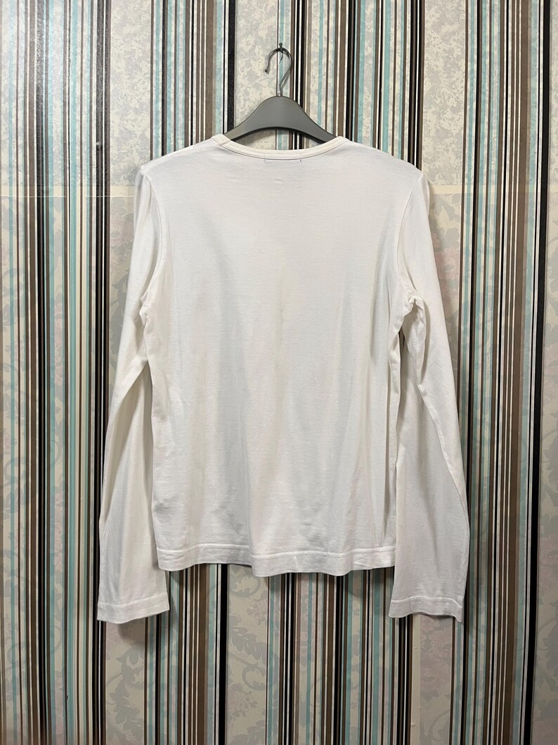 May include: A white long-sleeve shirt hanging on a black hanger. The shirt has a crew neck and is made of a soft, lightweight fabric. The sleeves are long and end at the wrists. The shirt is plain and simple.