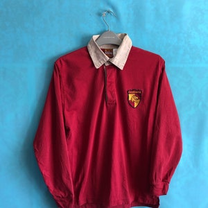 Peut inclure: Un maillot de rugby rouge à manches longues avec un col blanc et un blason représentant un lion sur un bouclier avec le texte "Gryffindor" en or.