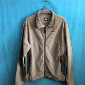 May include: A tan fleece jacket with a zipper closure and a black logo on the left chest. The jacket has two zippered pockets on the lower front and a black accent on the sleeves.