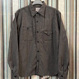 May include: A gray long-sleeve button-down shirt with two chest pockets and a small tag on the right pocket. The shirt has a herringbone pattern and a classic collar. The shirt is hanging on a metal hanger.
