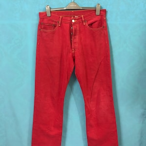 May include: Red denim jeans hanging on a hanger. The jeans have a button fly and are a straight leg style.