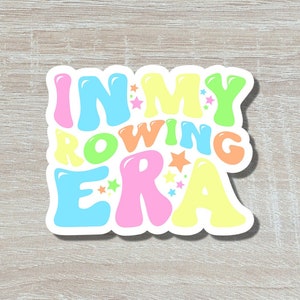 May include: A sticker with the text "In My Rowing Era" in colourful, bubble-like letters with stars. The sticker is on a light brown wood background.