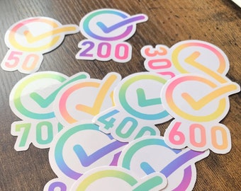 May include: A collection of rainbow-colored stickers with checkmarks and numbers. The stickers are arranged in a pile on a wooden surface. The numbers on the stickers include 50, 100, 200, 300, 400, 600, 700, 800, and 1K.