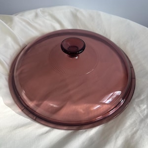 Peut inclure: Un couvercle rond en verre violet transparent avec une petite poignée circulaire. Le couvercle présente un motif texturé rayonnant depuis le centre. Le couvercle est probablement pour une casserole ou une poêle.