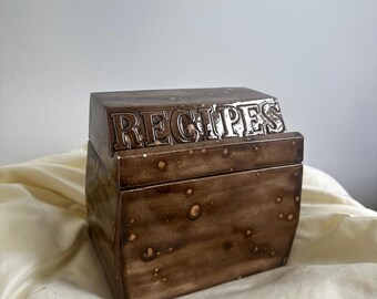 May include: A brown ceramic recipe box with the word "RECIPES" embossed on the lid. The box has a rectangular shape with a slightly angled lid and a speckled texture. The box is sitting on a cream-colored fabric.