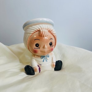 Puede incluir: Figura de cerámica vintage de un bebé sentado con un traje de marinero. La figura tiene un sombrero blanco con una raya azul, un traje blanco con un lazo azul y zapatos negros. El bebé tiene mejillas rosadas y una ceja ligeramente fruncida.