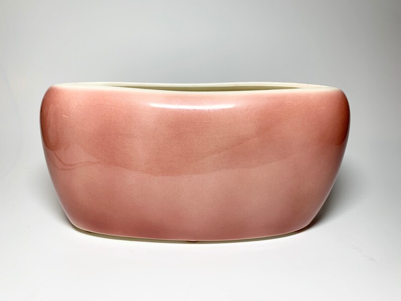 May include: A pale pink ceramic planter with a rounded, oval shape. The planter has a smooth, glossy finish.