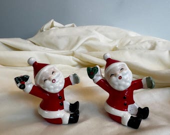 May include: Two vintage ceramic Santa Claus figurines with red suits, white trim, and black boots. Each Santa is holding a sprig of holly and has a smiling face.