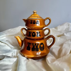 Puede incluir: Un juego de tetera de cerámica marrón con tapa y dos jarras más pequeñas. La tetera está decorada con letras negras que dicen "Tea for Two" con un pequeño corazón.