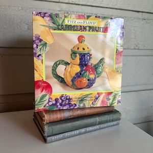 Könnte beinhalten: Ein Fitz and Floyd Caribbean Fruit Keramik-Simmertopf in der Originalverpackung. Die Schachtel zeigt eine farbenfrohe Abbildung der fruchtförmigen Teekanne mit Deckel, grünem Henkel und Ausguss, umgeben von gemalten Trauben, Birnen und Äpfeln.