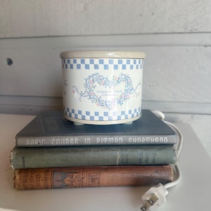 May include: A cylindrical electric potpourri crock by Rival with a cream-coloured base, featuring a blue and white checkered border at the top and bottom, and a central heart-shaped floral vine design with the text Potpourri Crock by RIVAL.