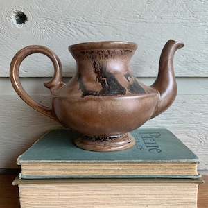 May include: A brown ceramic teapot with a dark brown glaze and a black design. The teapot has a rounded body and a spout on the side. It is sitting on top of two books.