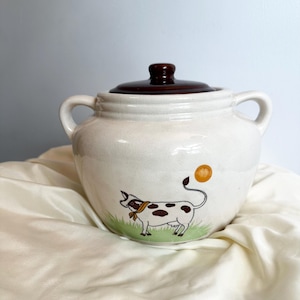 Puede incluir: Tarro de galletas de cerámica color crema con tapa y asas marrones. El tarro presenta una ilustración de vaca de dibujos animados con manchas marrones, una bufanda amarilla y una bola amarilla al final de su cola. El tarro está sobre una tela blanca.