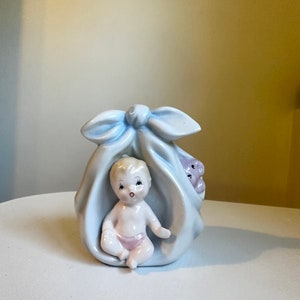 Puede incluir: Una figurita de porcelana de un bebé en una manta azul con un lazo. El bebé está sentado con un pañal rosa y un osito de peluche asomándose de la manta.