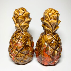 May include: A pair of brown ceramic pineapple salt and pepper shakers with orange and white accents.