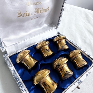 May include: A set of six vintage gold-coloured metal bottle stoppers in a decorative box. The box is white with a blue floral pattern and a blue satin interior. The box is labelled "Sainte Médard Paris".