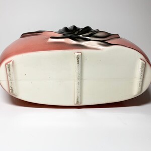 May include: The bottom of a pink ceramic vase with a white glaze. The vase has three raised feet on the bottom.