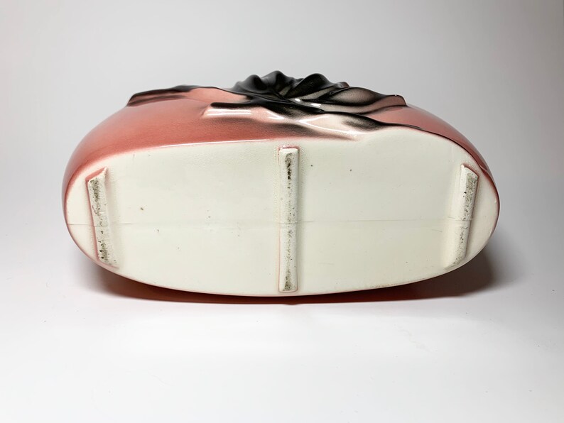 May include: The bottom of a pink ceramic vase with a white glaze. The vase has three raised feet on the bottom.