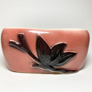 May include: A pink ceramic planter with a brown stylized leaf design.