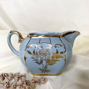 May include: A light blue creamer with gold trim and a floral design. The creamer has a scalloped edge and a handle.