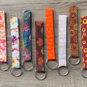 May include: A collection of eight fabric key fobs with various patterns. The patterns include floral, leaf, and abstract designs in colors like orange, pink, blue, and yellow. Each fob has a silver-colored key ring attached.