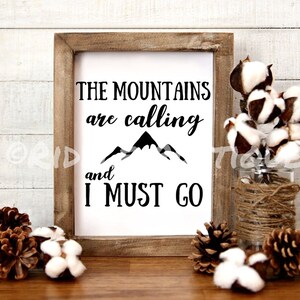 以下が含まれることがあります： 「The mountains are calling and I must go」という黒い文字が書かれた木製の額縁付きの看板。テキストの上にスタイリッシュな山脈のグラフィックがあります。看板は、松ぼっくりと綿のボールが置かれた木製の表面の上に置かれています。