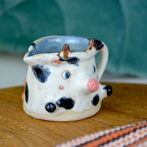 May include: A handcrafted ceramic cow creamer. The white creamer features black spots, a pink nose, and blue eyes. It has a small handle and a blue interior. The creamer is displayed on a wooden surface.