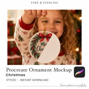 May include: A round white ceramic Christmas ornament featuring a printed green pine wreath with red berries and a red bow on the left side, while the right side remains blank. The ornament hangs from a gold beaded chain against a blurred background.