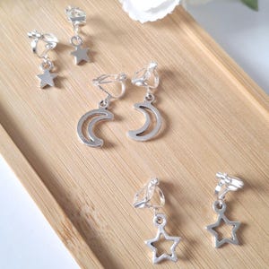 May include: Silver clip-on earrings featuring star and crescent moon designs. The earrings are presented on a light brown wooden tray, with white flowers in the background. The earrings are small and delicate.