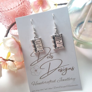May include: Silver earrings with a book charm that says "Once Upon A Time". The earrings are displayed on a white card with the text "Debs Designs Handcrafted Jewellery" and a website address.