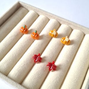 May include: A set of six stud earrings featuring miniature orange pumpkins with green stems and red maple leaves. The pumpkin earrings include two plain orange pumpkins and two pumpkins with small white flower accents on the front surface.