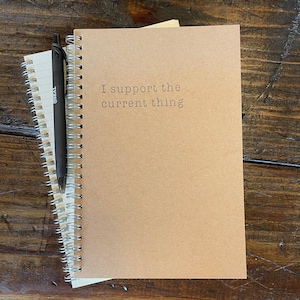 May include: A spiral-bound notebook with a kraft paper cover and the phrase "I support the current thing" printed in a simple font. A black pen rests on top of the notebook, which is layered over a smaller, white spiral notebook.