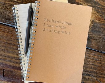 May include: A spiral-bound notebook with the text "Brilliant ideas I had while drinking wine" printed on the cover. A black pen rests on top of the notebook, which is sitting on a wooden surface.