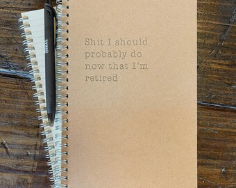 May include: A spiral-bound notebook with the text "Shit I should probably do now that I'm retired" printed on the cover. A black pen rests on top of the notebook, which is next to a smaller notepad. The notebook is tan.