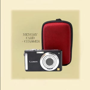 Könnte beinhalten: Eine schwarze Panasonic Lumix Digitalkamera mit silbernem Objektivring und einer roten, strukturierten Tragetasche. Der Text MEMORY CARD + CHARGER ist links neben der Kamera zu sehen. Die Kamera verfügt über ein Leica-Objektiv und silberne Akzente.