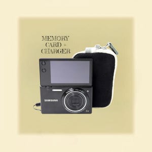 Op de afbeelding: Een zwarte Samsung digitale camera met een opklapbaar scherm en een Schneider-Kreuznach lens, vergezeld van een zwart-witte tas met ritssluiting. De tekst "MEMORY CARD + CHARGER" staat in een donker schreeflettertype boven de camera.