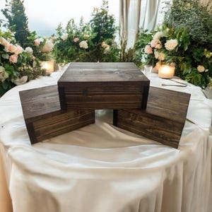 May include: Three dark stained wooden boxes of varying sizes are arranged on a white tablecloth. The boxes have a square shape and are made of wood with visible grain. The background features greenery and soft lighting, suggesting a wedding or event setting.