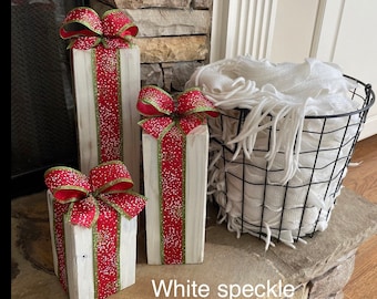 Puede incluir: Tres bloques de madera rectangulares pintados de blanco con acabado desgastado, cada uno envuelto en una cinta roja con motas blancas y bordes verdes, rematado con un gran lazo rojo. Una cesta de alambre negro contiene una manta blanca con flecos.