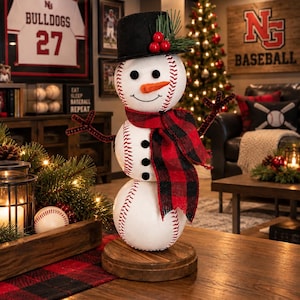 May include: A snowman figurine constructed from three white baseballs with red stitching, wearing a black top hat with green pine needles and red berries. It features a carrot-colored nose, black button eyes, and a red and black buffalo plaid scarf.