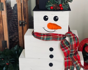 Puede incluir: Un muñeco de nieve decorativo hecho de tres bloques de madera blancos apilados. Tiene un sombrero de copa negro adornado con agujas de pino, una pequeña piña y bayas rojas. La cara tiene ojos negros, una nariz de zanahoria de fieltro naranja.