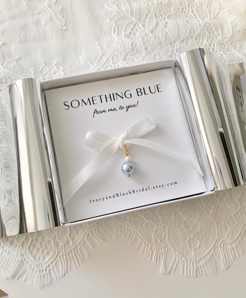 May include: A small charm featuring a light blue bead and a tiny white pearl suspended from a white ribbon bow, presented in a white box with the text SOMETHING BLUE from me, to you! and IvoryandBlushBridal.etsy.com printed on the card insert.